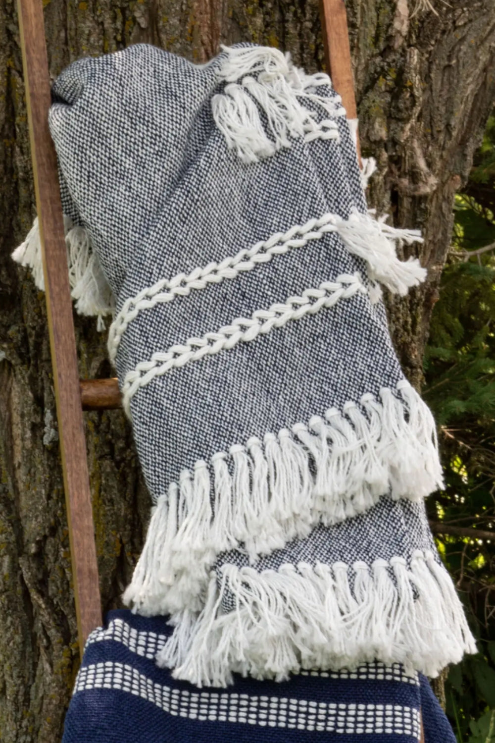 Gray woven blanket with white fringe draped over a wooden ladder against a tree.